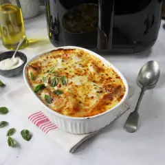 En gyllene lasagne i airfryer, toppad med smält ost och färsk basilika. Serveras i en vit ugnsform med en sked bredvid.