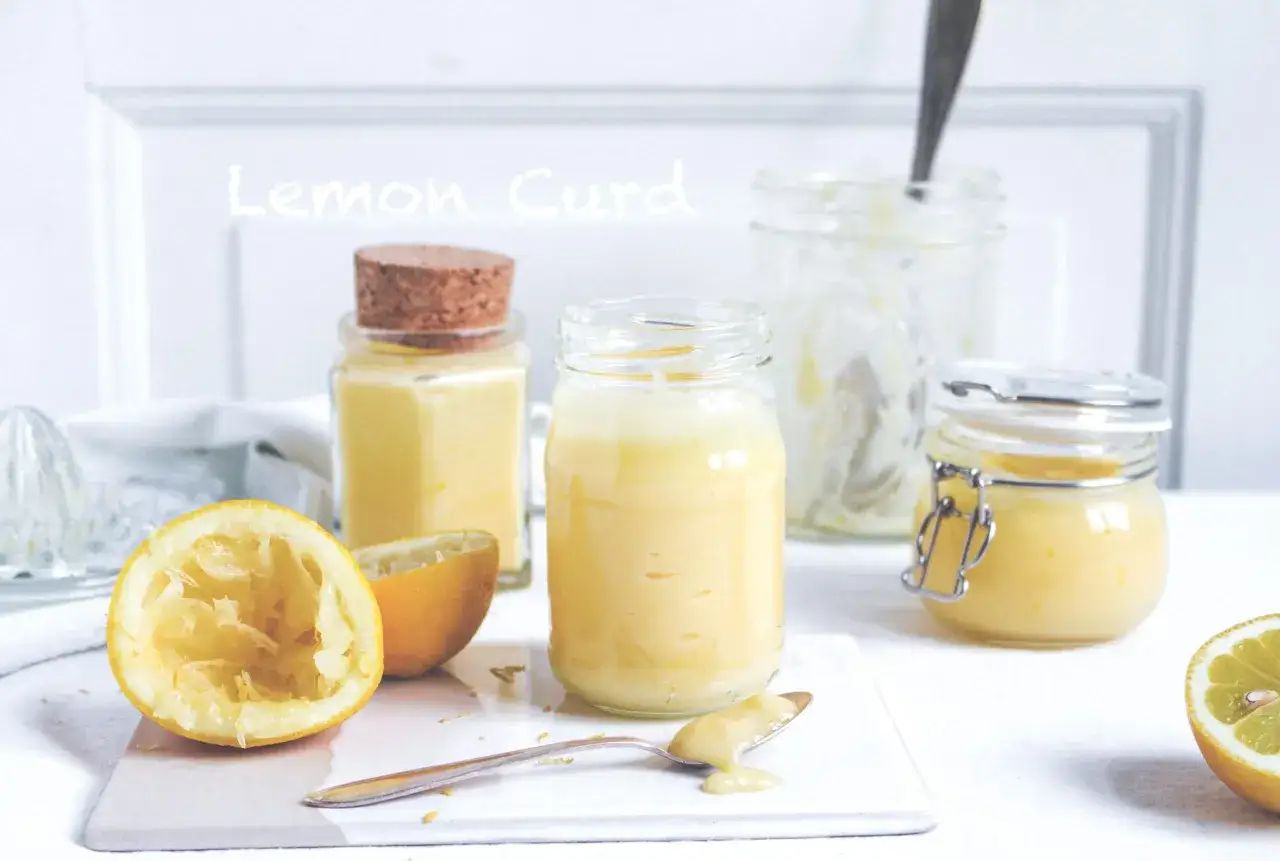 Burkar med hemgjord lemoncurd, en pressad citron och en sked med kräm. Perfekt för ditt lemoncurd recept!