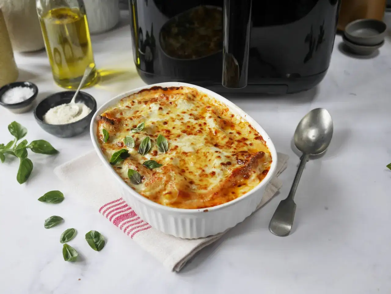 En gyllene lasagne i en vit form, nygjord i airfryer. Serverad med färsk basilika och en sked redo.