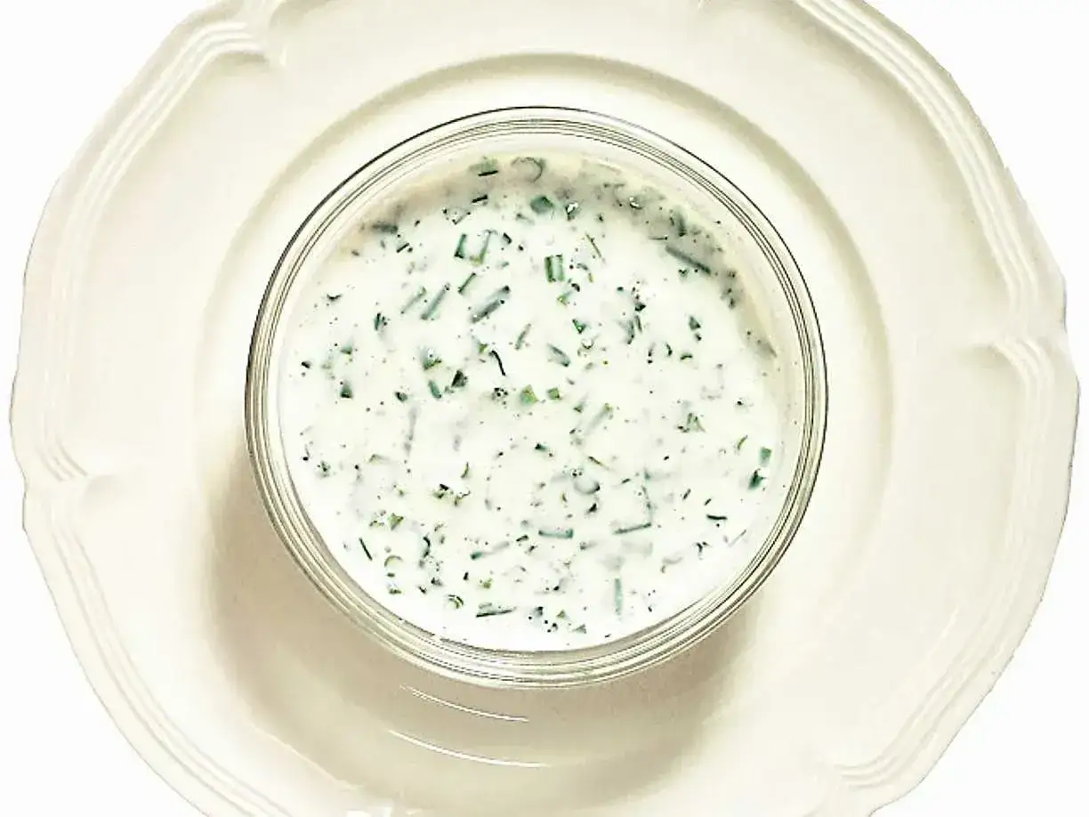 En skål med krämig creme fraiche sås med finhackade örter, serverad på en vit tallrik. Perfekt till sommarmaten!