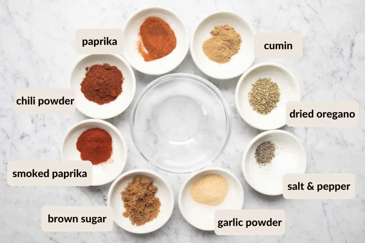 Små skålar med kryddor som paprika, chili, spiskummin, oregano, salt, peppar, farinsocker och vitlökspulver arrangerade runt en tom skål. Ingredienserna för en härlig cajun krydda.